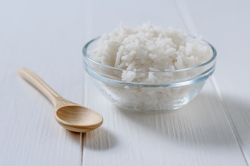 Glass bowl with long grain rice and wooden spoon on white wooden table.