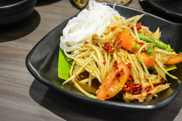 famous and popular Thai street food, green papaya spicy salad with pickled fish or Som Tum served with Thai rice noodles in black plate on wooden table background
