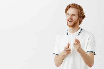 Hey how you doing. Portrait of charming flirty and confident happy redhead guy with beard in white polo shirt pointing at camera with finger guns and winking with broad macho smile over gray wall