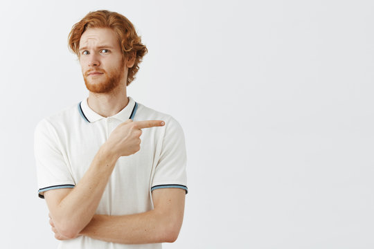 Doubtful and surprised redhead young businessman in white plo shirt frowning looking intense and unsure while pointing right and tilting left with uncertain look being unimpressed and displeased