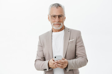 Portrait of stylish and smart confident senior businessman in glasses and elegant formal suit holding smartphone against chest smirking at camera pleased with new useful features of gadget