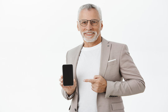 Delighted friendly handsome male with white beard and moustache in elegant suit and black frames holding cellphone pointing at smartphone screen showing image in gadget, smiling happily