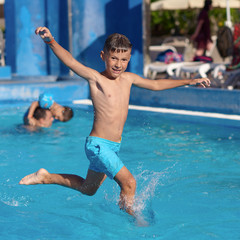 Caucasian boy having fun making fantastic jump into swimming pool at resort. His arms and legs are wide open. He is smiling and looking towards the camera.