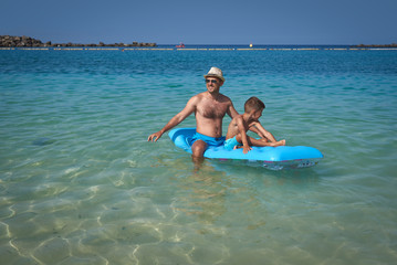 Obraz premium Attractive European man with his son is sitting on a blue air mattress on the beach. They are relaxing and enjoying their holidays.