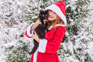 Christmas time. Portrait of beautiful young Santa christmas woman posing wearing santa claus dress outdoor with black cat. Snowflakes are flying around 