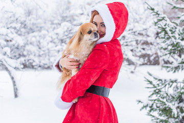 Christmas time. Portrait of beautiful young Santa christmas woman posing wearing santa claus dress outdoor with little dog. Snowflakes are flying around  