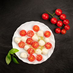 A photo of a plate of Italian Caprese salad with Mozzarella cheese, cherry tomatoes, and basil leaves, shot from the top on a dark background with copy space