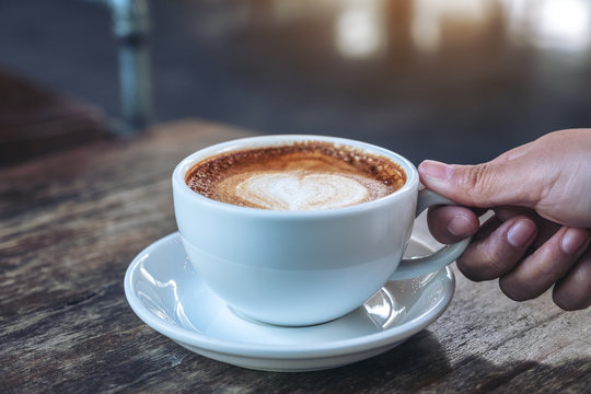 Closeup image of a hand holding a blue cup of hot coffee on wooden table in cafe