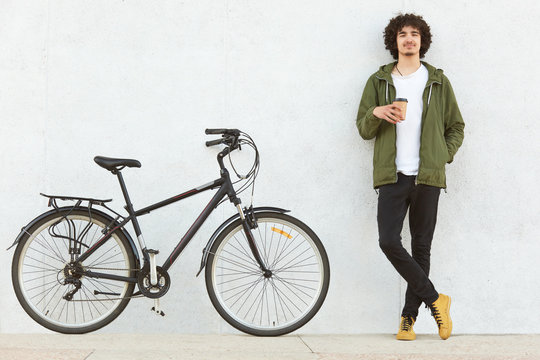 Photo of attractive curly man dressed in fashionable clothes, has coffee break after riding sport bicycle, models over white concrete wall, enjoys nice trip with friend. Youth, transportation concept
