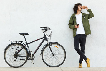 Horizontal shot of fashionable youngster keeps hands crossed, holds mobile phone, sends text...