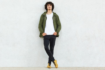 Full length shot of attractive self confident curly man dressed in green anorak, keeps both hands in pockets, enjoys spare time, isolated over white studio wall. People, youth and fashion concept - obrazy, fototapety, plakaty