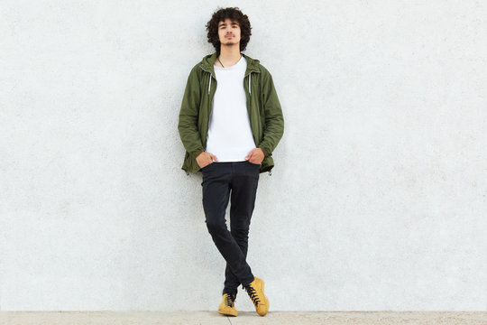 Full length shot of attractive self confident curly man dressed in green anorak, keeps both hands in pockets, enjoys spare time, isolated over white studio wall. People, youth and fashion concept