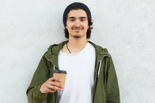 Headshot of pleased hipster guy holds takeaway coffee, dressed in white t shirt and green jacket, enjoys spare time, wears black hat, models over white background, has curly hair. People and drinking