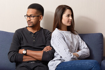 Photo of two offended young people in love feel jealous, turn from each other, keep hands crossed, have crisis in their family, pose at sofa, being displeased. Interracial relationship concept