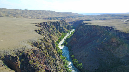 A blue river runs along the canyon. On the banks of the river grows green grass and trees. Boiling river in the canyon. Around the desert. View of the road. Steep edges of the canyon.