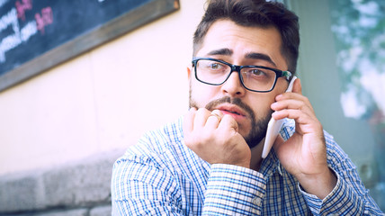 Young man in plaid shirt and eyeglasses has phone conversation, sitting at cafe.