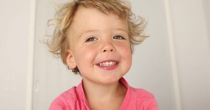 Adorable happy baby boy looks around and then smiles and laughs. 2 year old blond haired child looking at camera at home light background