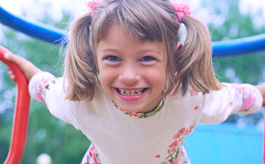 Portrait of cute Caucasian litte girl wearing white dress with flowers hanging on monkey bars on a summer day. Girl looking at camera smiling. Green leaves a seen in the background.