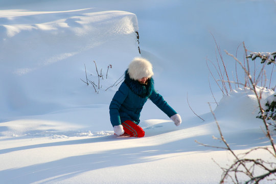 A lone traveler, a girl in a white fluffy hat and red pants, wearing a blue jacket on a clear winter day, goes through the snowdrifts, knee-deep in deep snow falls, hardly makes her way to the goal.