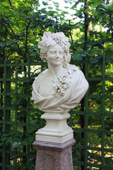 Fototapeta premium Antique Sculpture of Woman Head and Bust Representing Autumn in Summer Gardens. Historical White Statue Outdoors on Green Leaves Background at the City Park