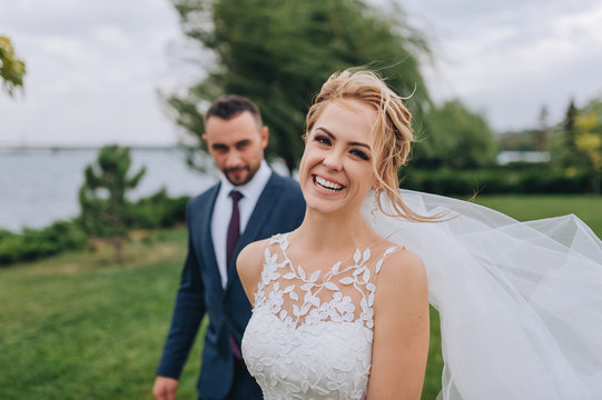 Blonde and smiling bride and bearded groom walk, holding hands, in the park with green grass and willows. Wedding portrait of beautiful newlyweds. Wedding photography.