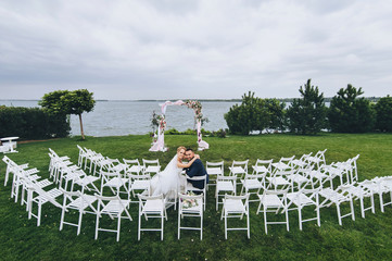 Beautiful newlyweds sit on chairs, against the backdrop of the arch and the sea. Wedding ceremony. The bearded bride and groom sit on a background of nature. Wide angle photo.