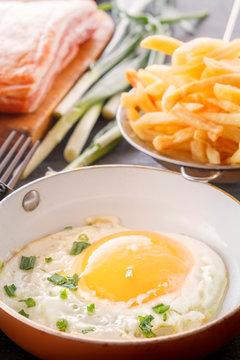 Fried egg in a pan, bacon, french fries and green onions are cooked for breakfast on a wooden gray table. Close-up