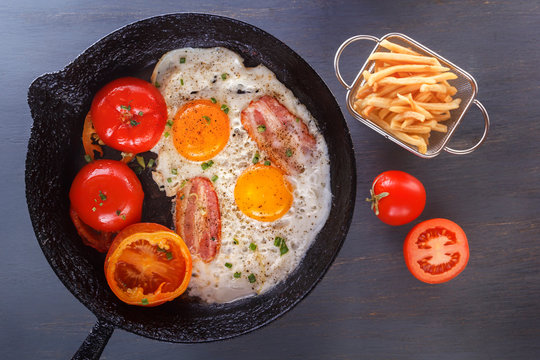 Fried eggs with bacon and tomatoes on an old cast-iron pan with french fries on a gray table. Close-up. Top view