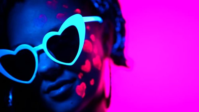 Portrait of fashion model woman with heart shaped glasses in neon light. Fluorescent unusual makeup glowing under UV light. Night club, party, halloween psychedelic concepts