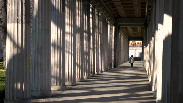 Rear view of young woman teenage girl mixed race female wearing camouflage jacket, walking through columns passageway during the day with a train going past, Berlin, Germany