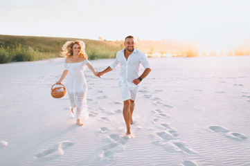 A bearded man and a blond woman run and hold hands on the white sand at sunset. Love in the desert newlyweds. The love story of fun and love people.