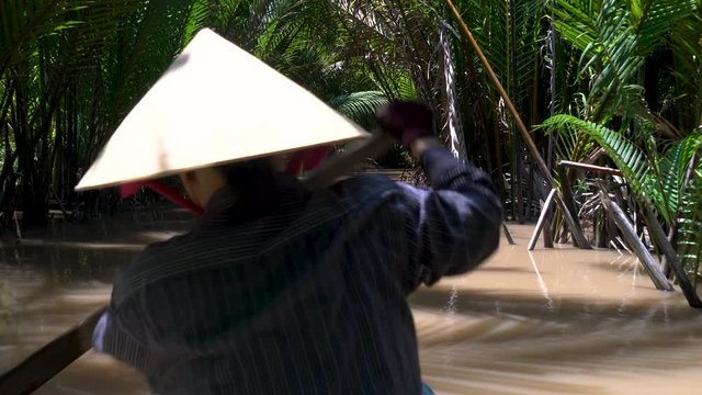 Rear view of a local Vietnamese woman wearing a leaf hat and paddling a traditional boat or canoe in the Mekong Delta, Vietnam