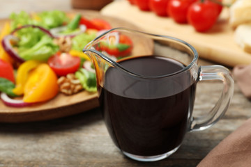 Balsamic vinegar in glass dish near plate with vegetable salad on wooden table, closeup