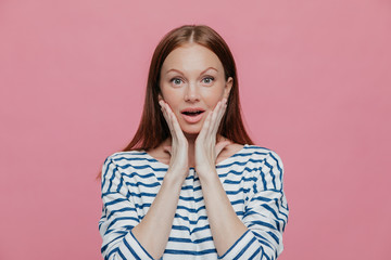 Beautiful dark haired amazed woman touches cheeks with both hands, dressed in striped clothes, looks surprisingly at camera, poses against pink background. People, reaction and emotions concept