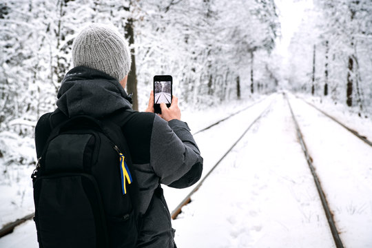 A guy with a backpack photographs on the phone the tram rails running through the snowy forest.
