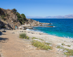 Two girl friends walking on rocky seashore next to Bataria white sand beach at Kassiopi Bay. Blue sea and clear sky, summer Greece, corfu