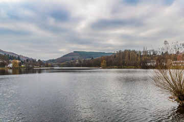 Obraz premium Landscape of Doyards lake surrounded by autumnal trees and wild plants, Vielsalm village in background against cloud-capped sky, light air over water surface, autumn day in the Belgian Ardennes