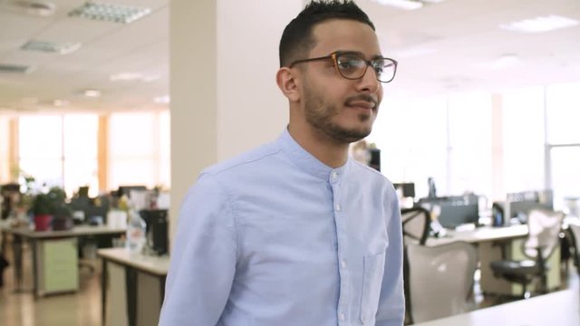 Medium shot of young bearded Asian man in glasses walking through office while colleagues working in the background