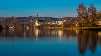 Landscape of Doyards lake with reflection on water surface, Vielsalm village, its St Gengoul church and wooded mountain against blue sky in background, cold winter evening in the Belgian Ardennes