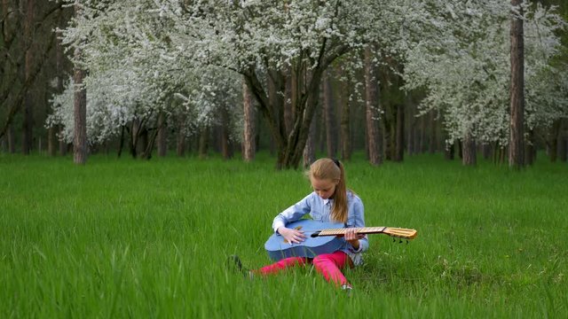 nice little girl guitarist playing solo. sitting in a wonderful place tune your guitar on background of a blossoming tree green grass in the spring forest