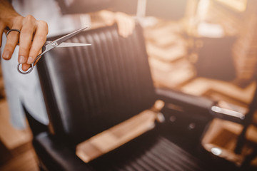 Close up. Men hairdresser holding scissors in hand against vintage-inspired black leather seats, back warmer overexposure