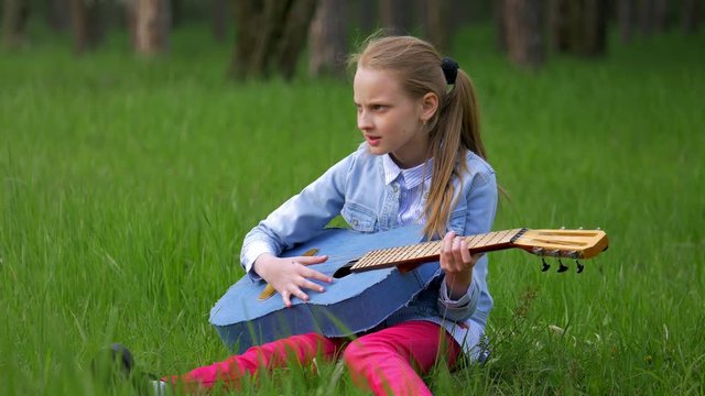 little girl in a jeans jacket having fun and play guitar at springtime sitting in green grass against the forest