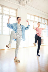 Two young dancers lifting one leg and arms while repeating move after trainer during workout in gym