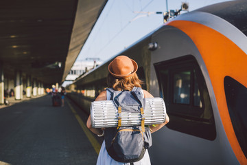 Theme transportation and travel. young caucasian woman standing at train station platform near train backs train background with backpack travel mat sleepy weather in dress and hat in summer
