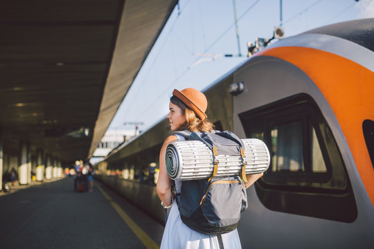Theme Transportation And Travel. Young Caucasian Woman Standing At Train Station Platform Near Train Backs Train Background With Backpack Travel Mat Sleepy Weather In Dress And Hat In Summer