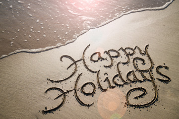 Happy Holidays message handwritten in smooth sand with an oncoming wave in the lens flare of the...
