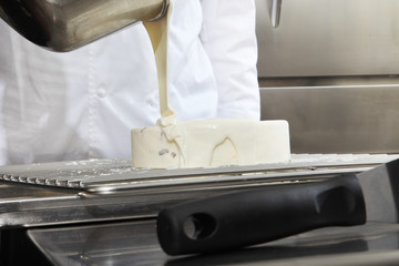 hands pastry chef prepares a cake, cover pouring white icing, working on a stainless steel industrial kitchen work top