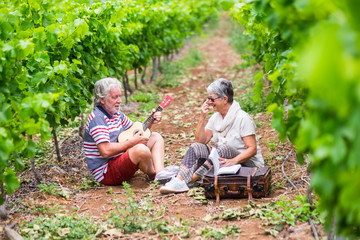couple of alternative aged older stay sitting down in a vineyard with the luggage and playing an ukulele acoustic guitar and reading a paper book. easy lifestyle in outdoor places and nature feel