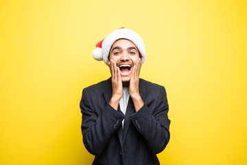 Closeup portrait of shocked and surprised handsome young man wearing red santa claus hat, hands in air, mouth eyes wide open, isolated on yellow background. Positive human emotion facial expression