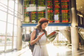 theme travel and transportation. Beautiful young caucasian woman in dress and backpack standing inside train station terminal looking at electronic scoreboard holding phone, map paper hand navigation © Elizaveta